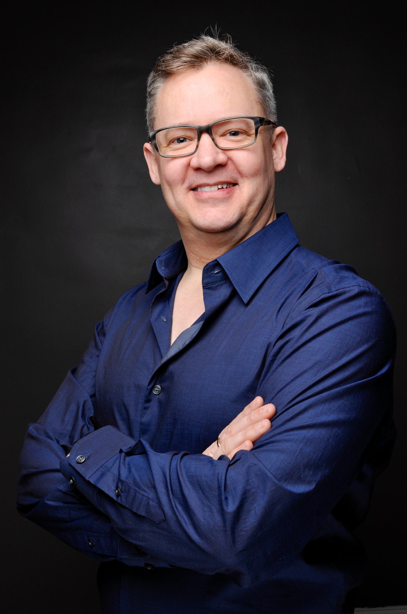 Smiling middle-aged man with glasses wearing navy blue shirt with arms crossed in studio.