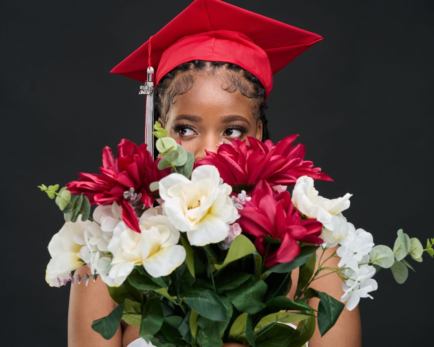 Young woman in red graduation cap holding bouquet of red and white flowers in front of her face.