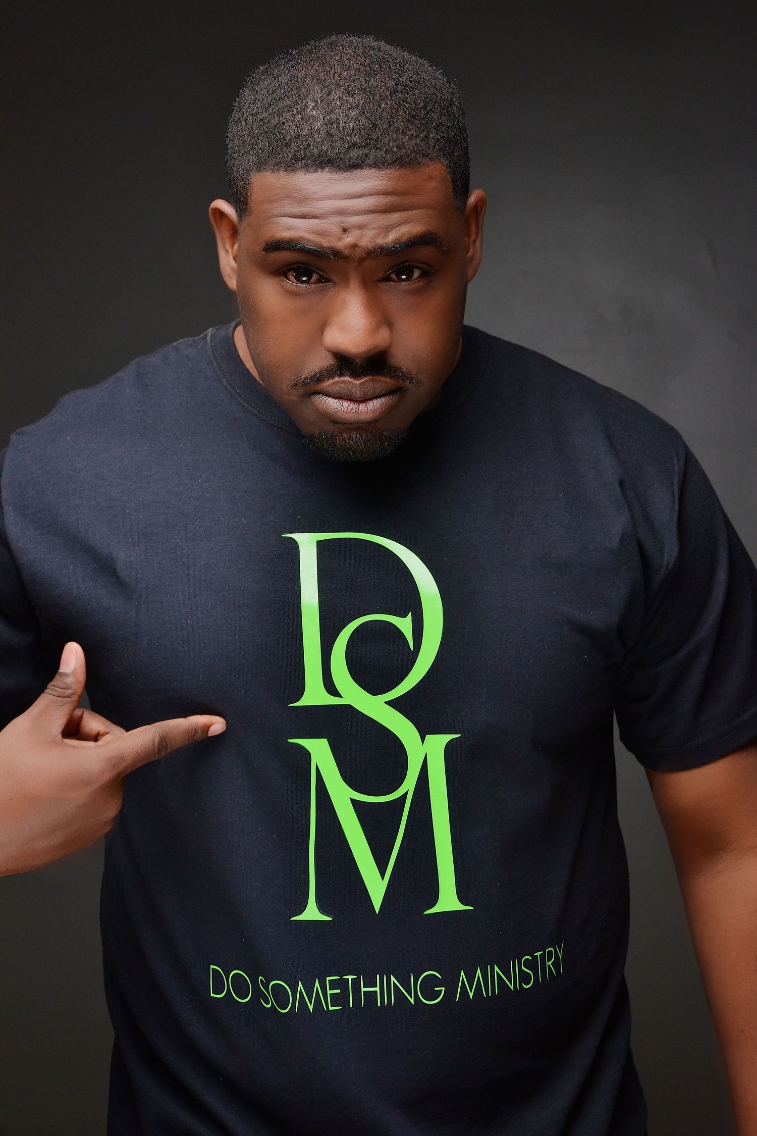 Man in black Do Something Ministry t-shirt pointing to logo against dark background.