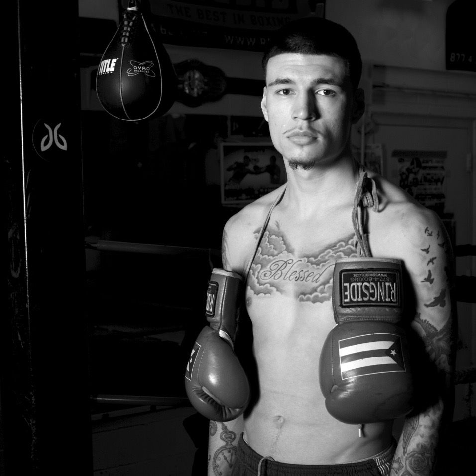 Black and white photo of a tattooed male boxer holding gloves in a gym near a speed bag.