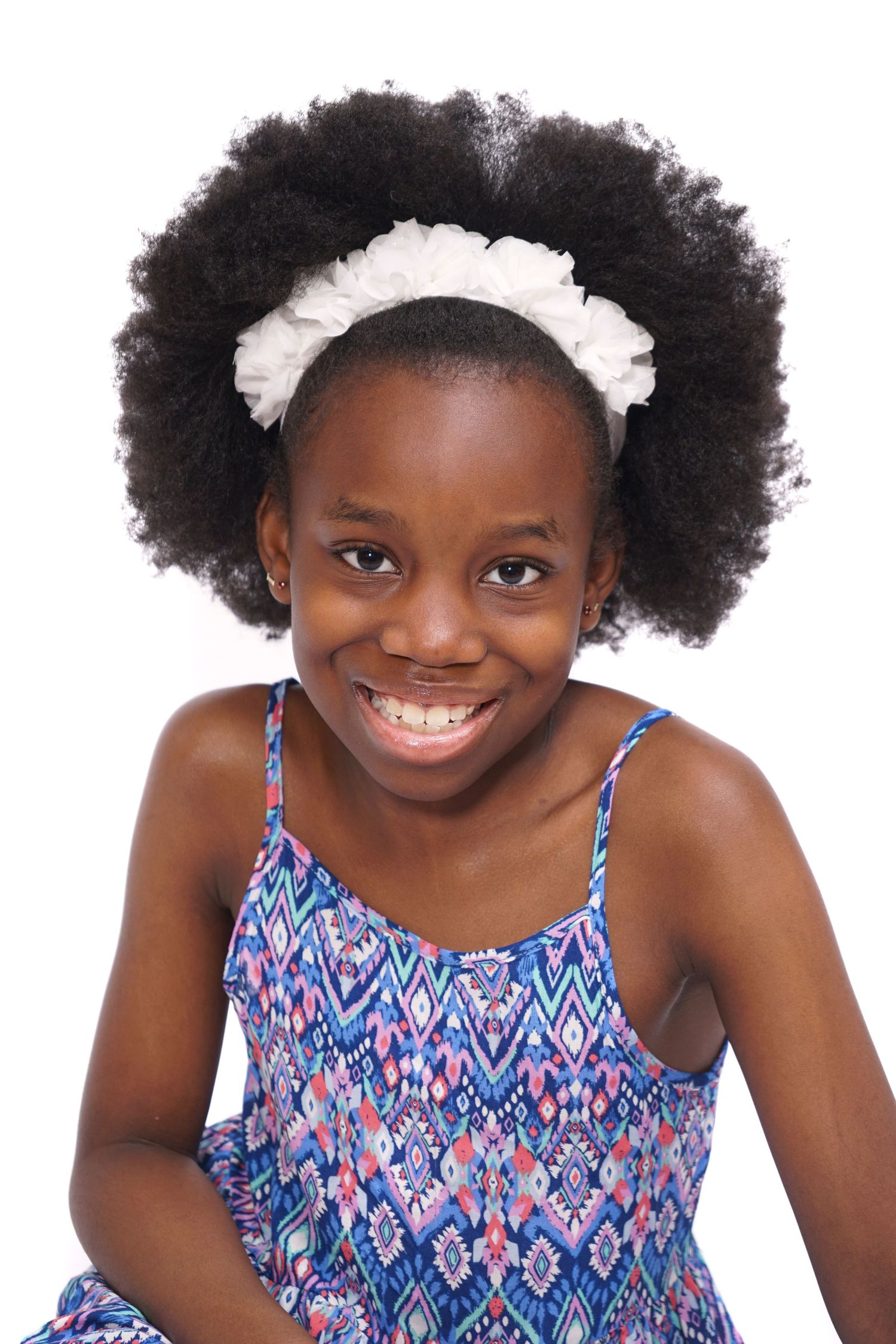 Smiling young Black girl with afro hair and white headband wearing a colorful blue sundress.