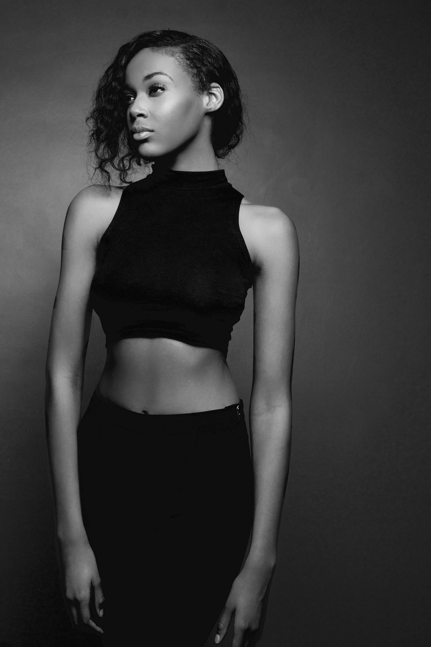 Slender young woman in black crop top and skirt poses in dramatic black and white studio portrait.