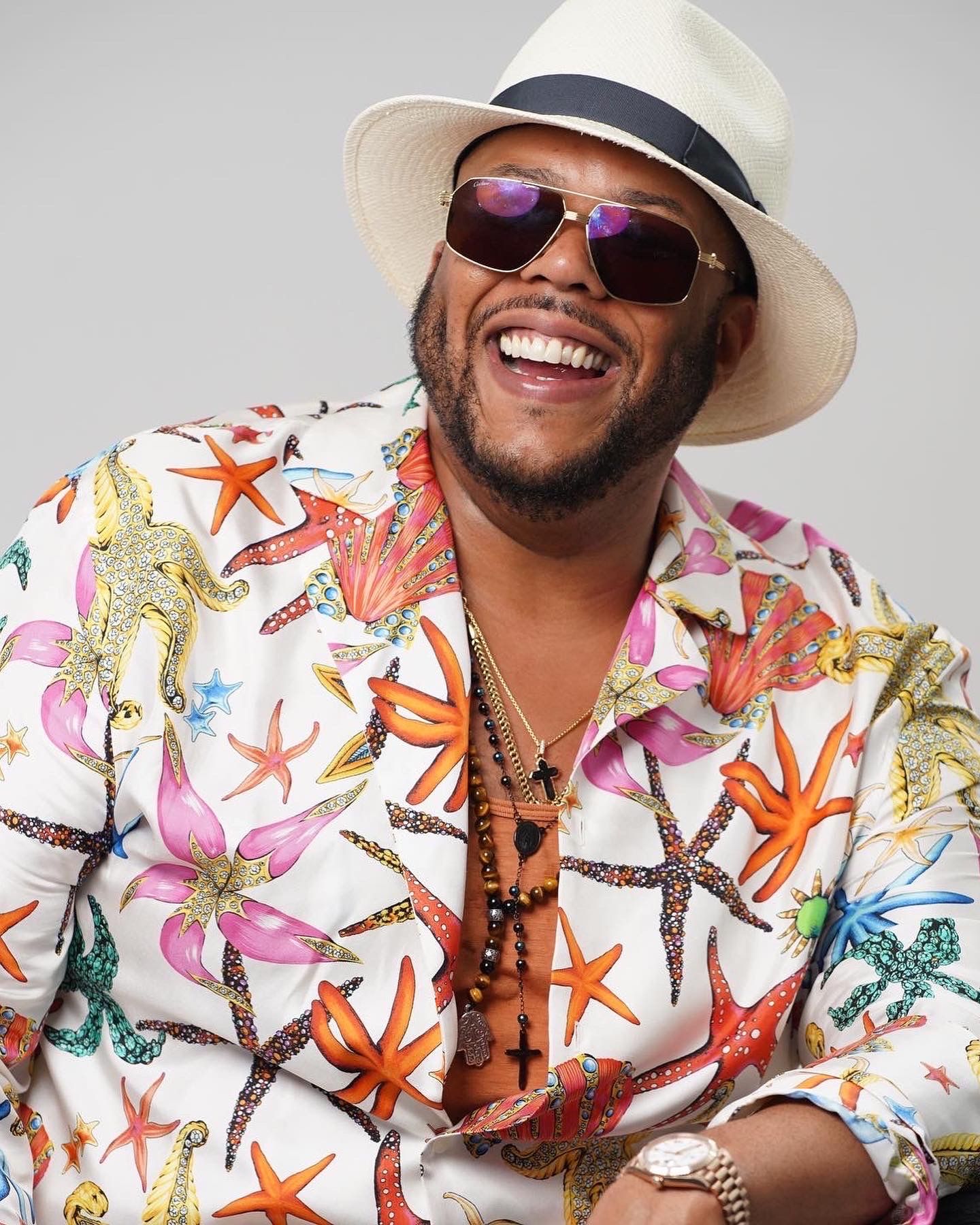 Man smiling in colorful tropical print shirt, white fedora hat, and sunglasses.