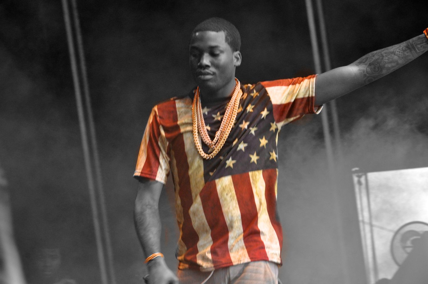 Male performer on stage wearing American flag t-shirt and gold chain necklace with arm raised to crowd.