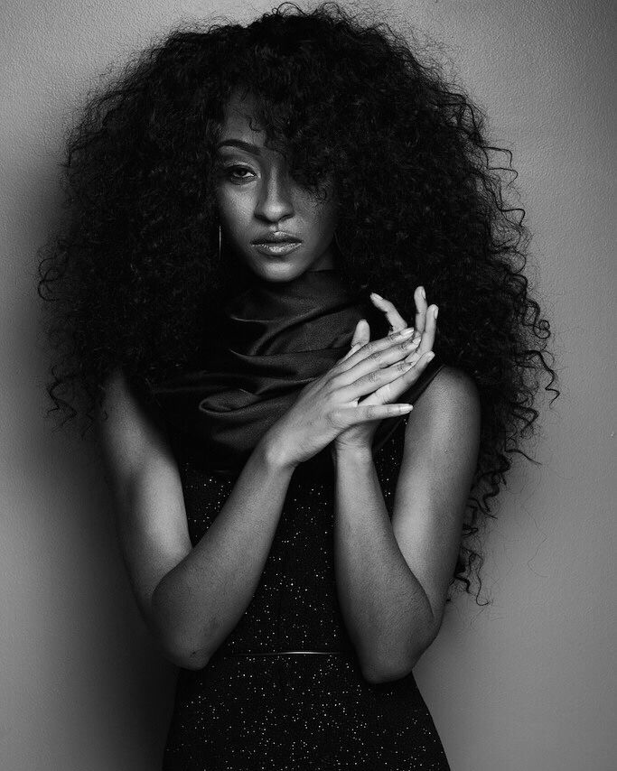 Black and white portrait of a woman with voluminous curly hair holding hands to her chest.