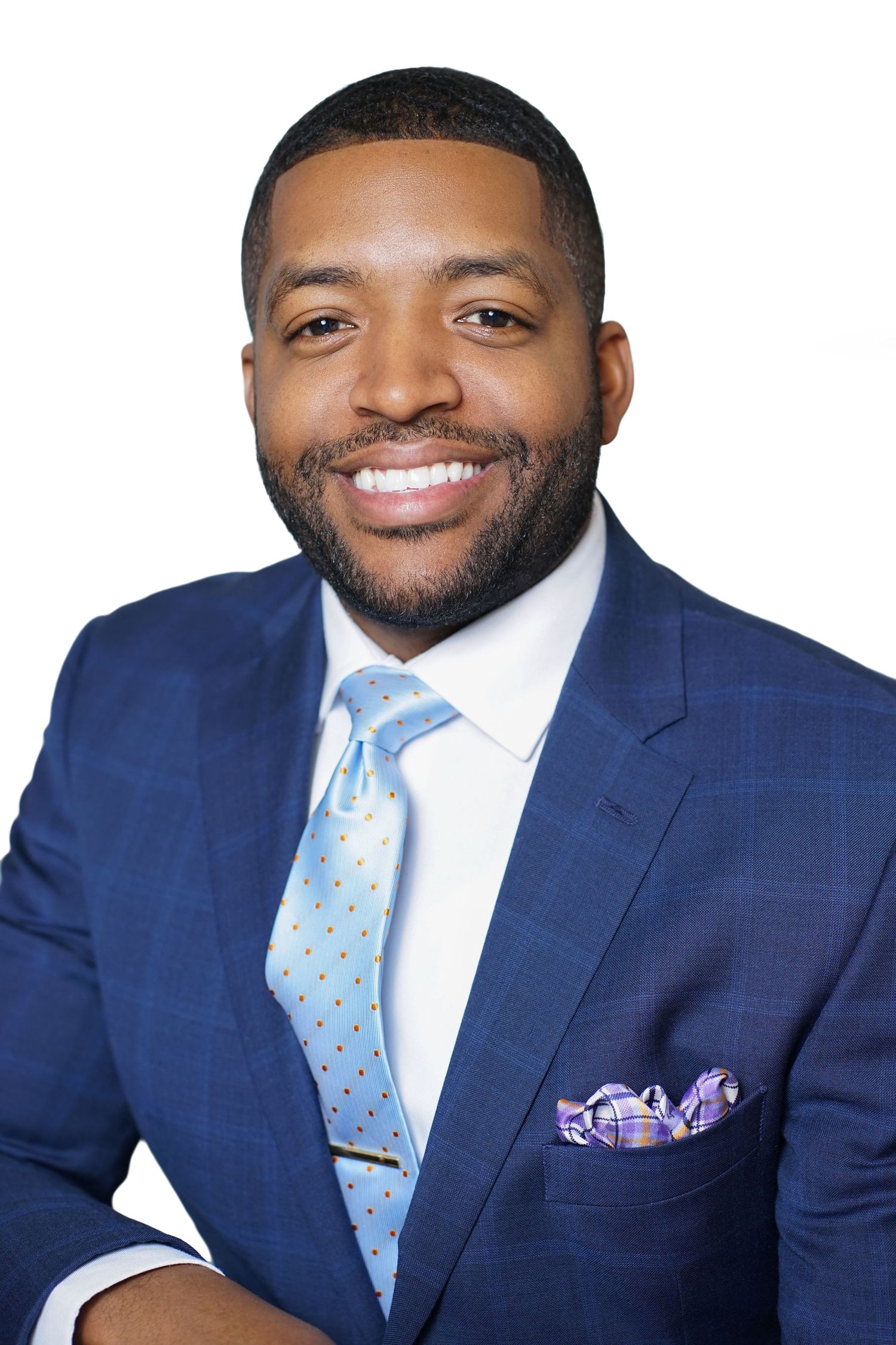 Smiling Black man in navy blue suit with light blue tie and pocket square, professional headshot on white background.