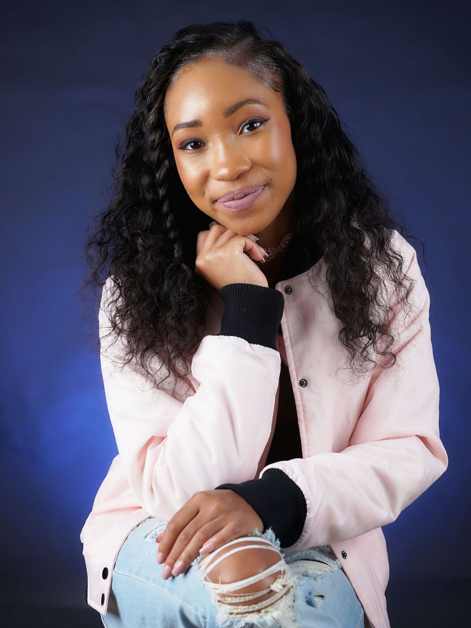 Young Black woman with long curly hair wearing pink jacket and ripped jeans posing on blue background.
