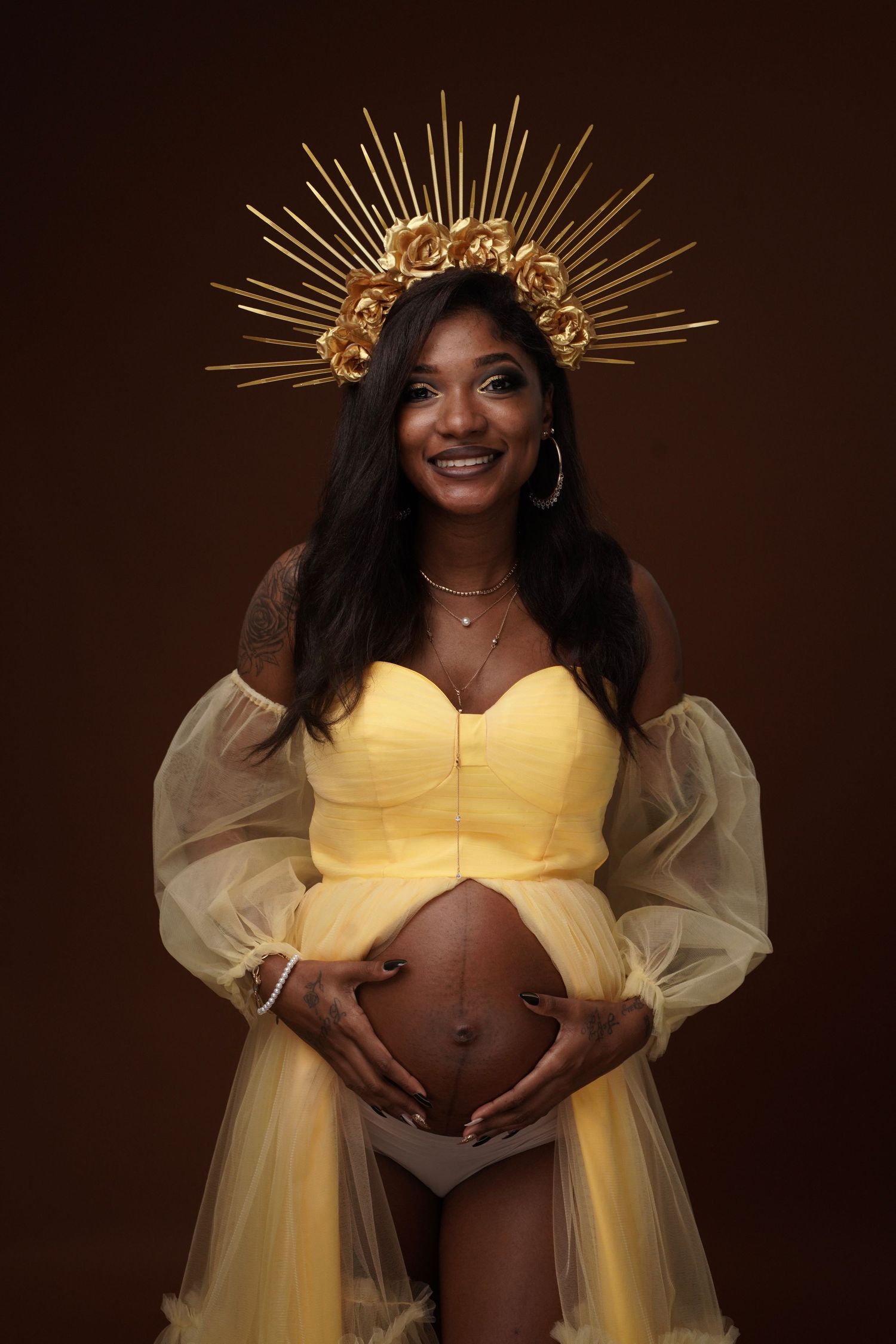 Smiling pregnant woman in yellow dress with gold crown headpiece cradling baby bump on dark background.