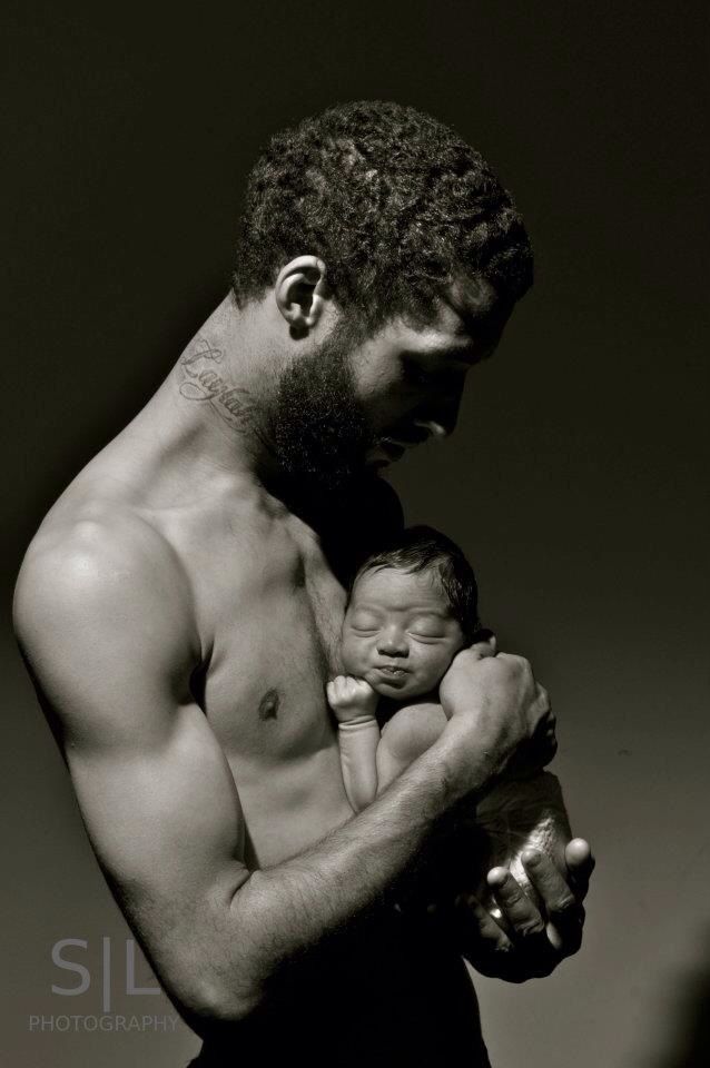 Black and white photo of a muscular shirtless man tenderly cradling a sleeping newborn baby against his chest.