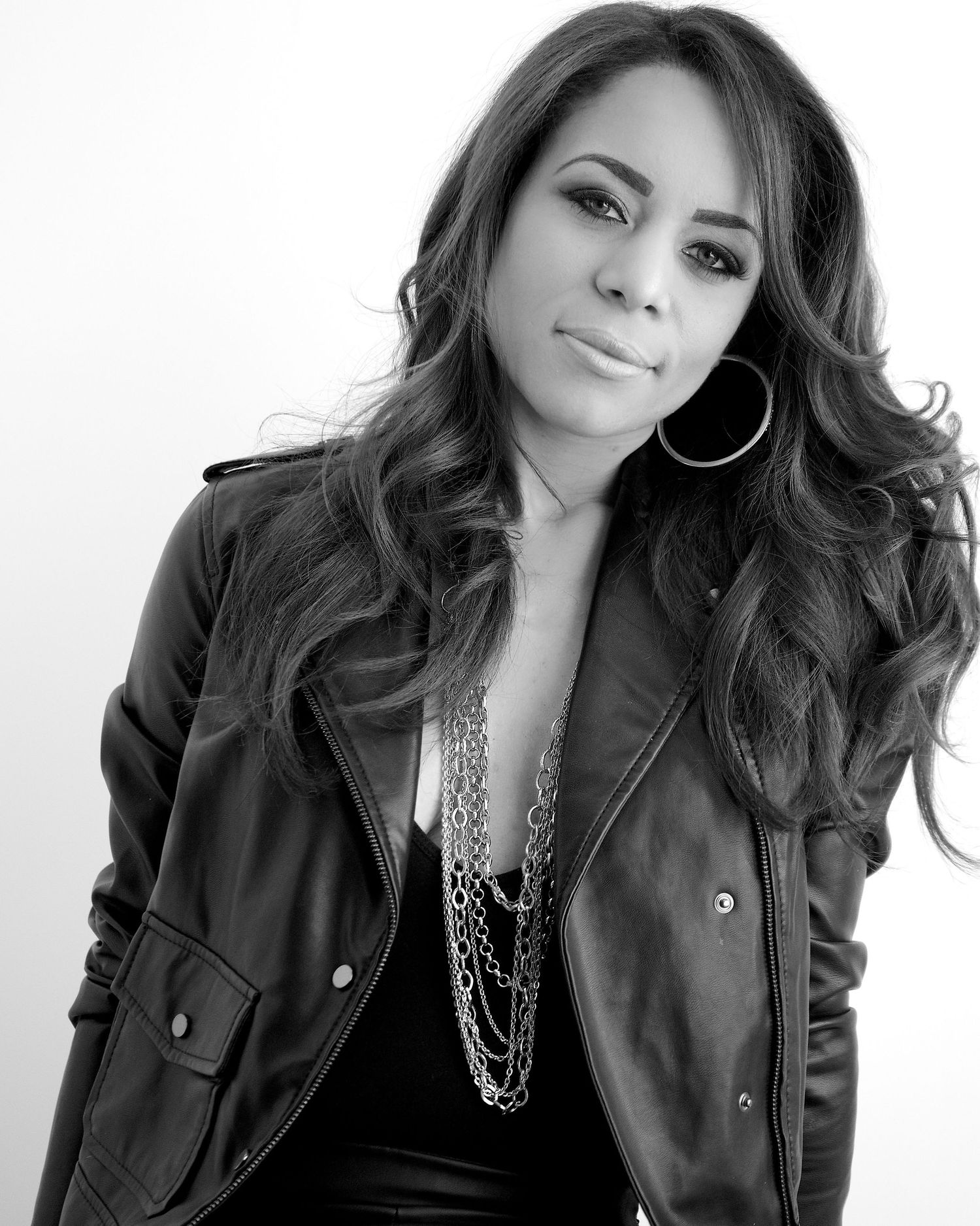 Black and white portrait of a woman in a leather jacket with layered necklaces and hoop earrings.