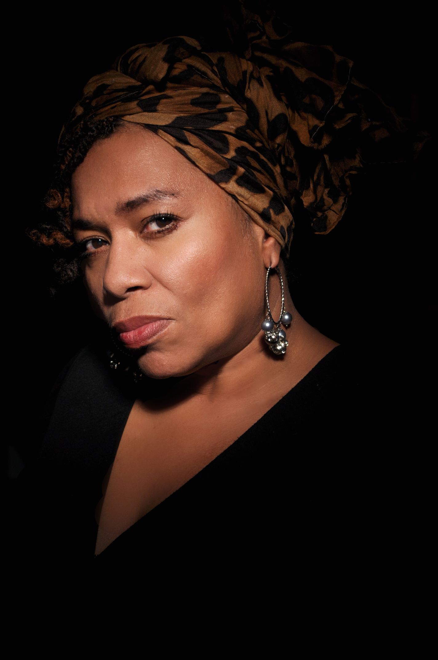 Woman wearing leopard print head wrap and silver earrings against dark background.