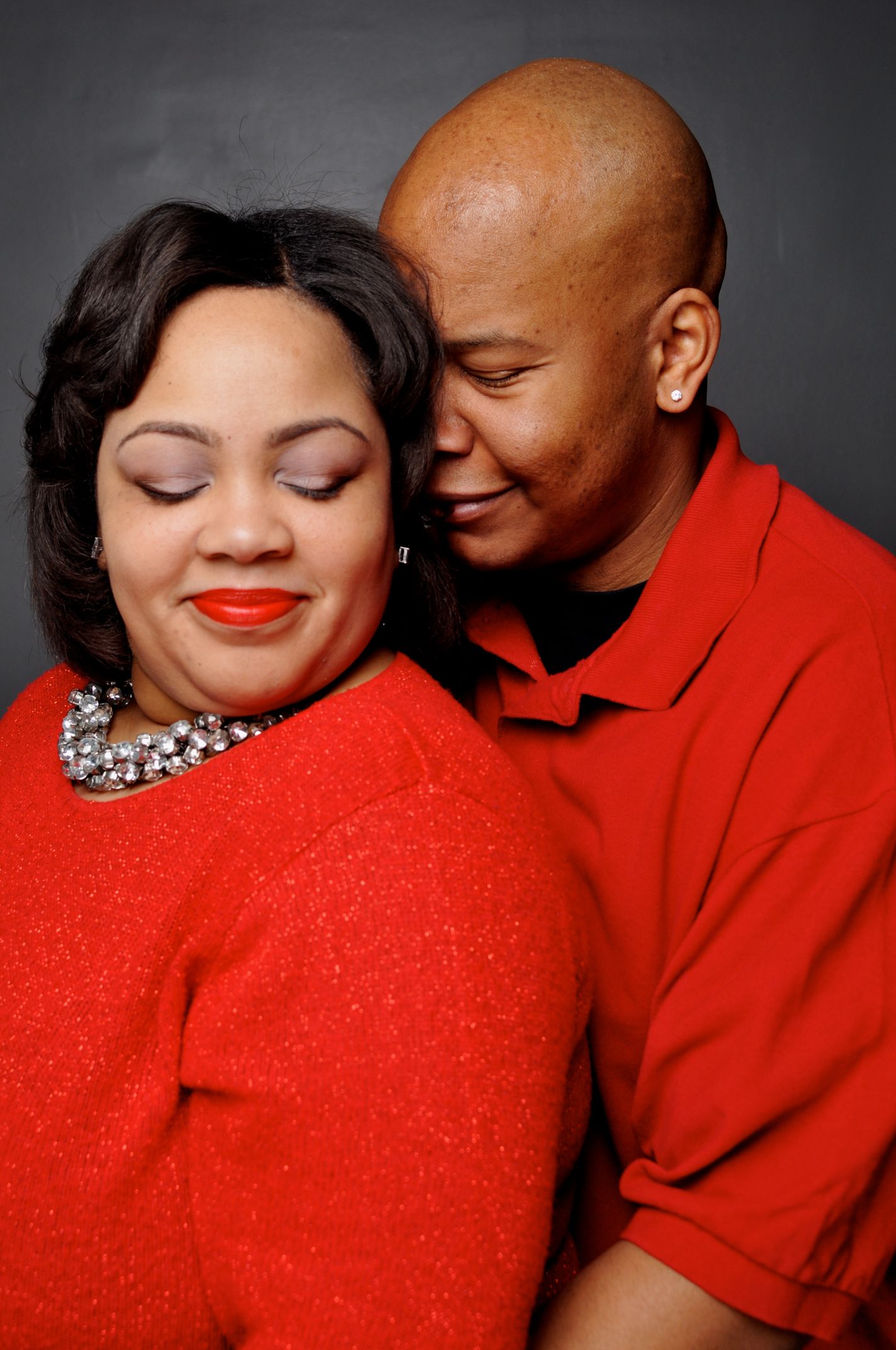A couple in matching red outfits share a tender moment, the man resting his head against the woman's cheek.
