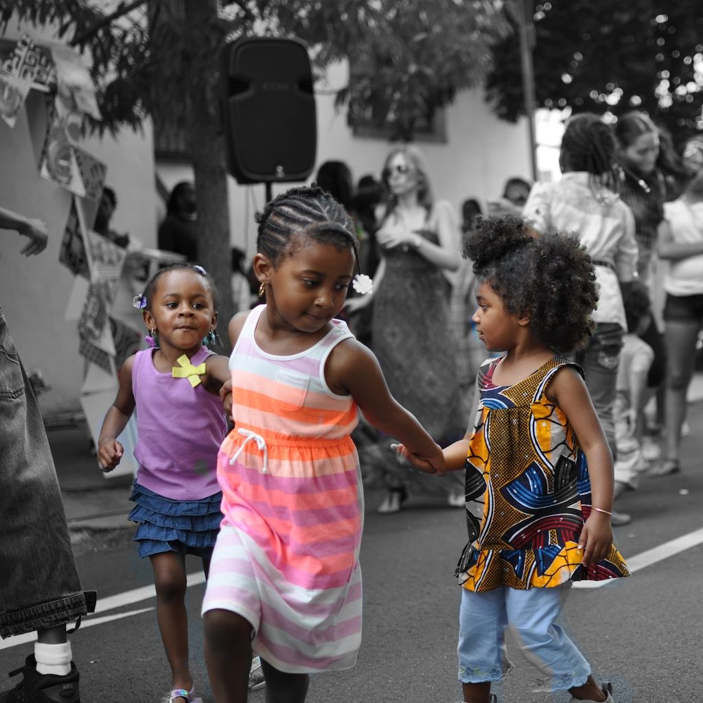 Three young Black girls holding hands and dancing together at an outdoor community event.