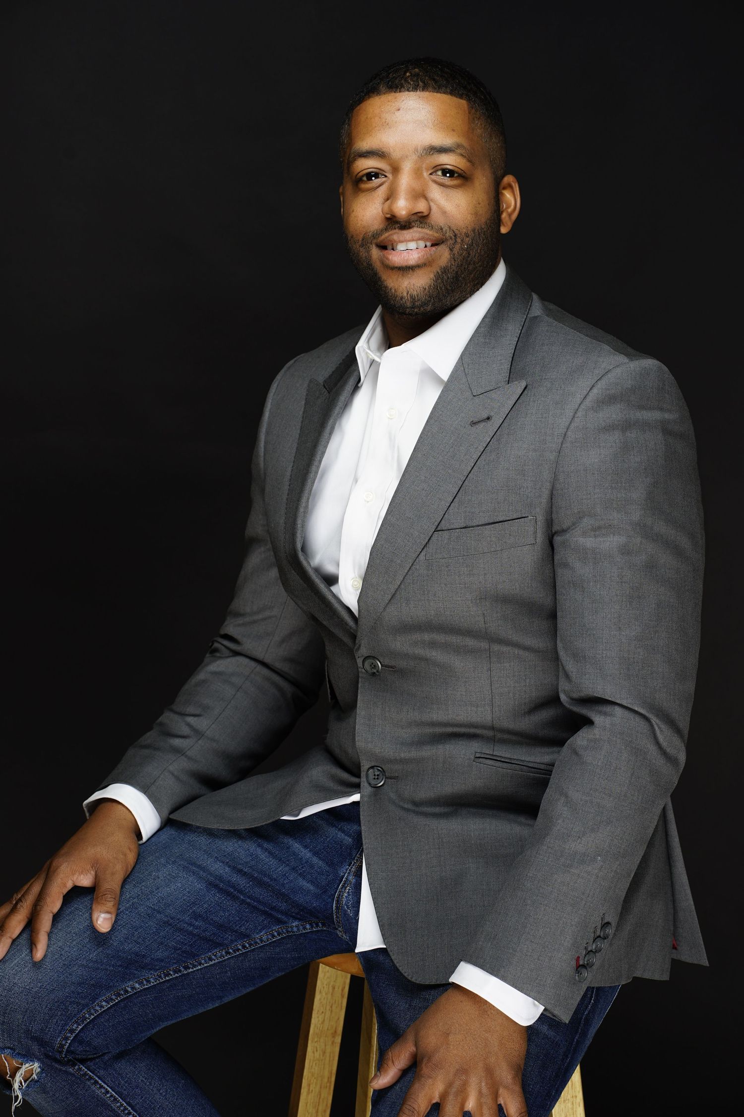 A smiling Black man in a gray blazer and jeans sits confidently on a stool against a dark background.