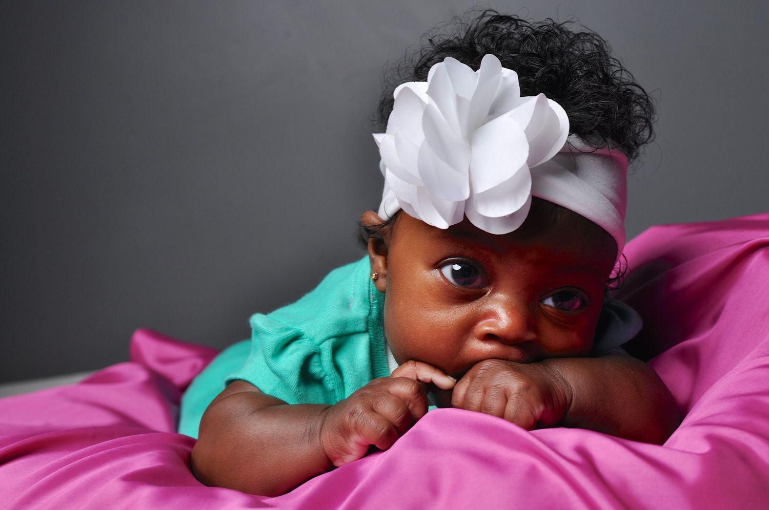 Adorable Black baby girl in teal outfit with white flower headband lying on pink fabric.