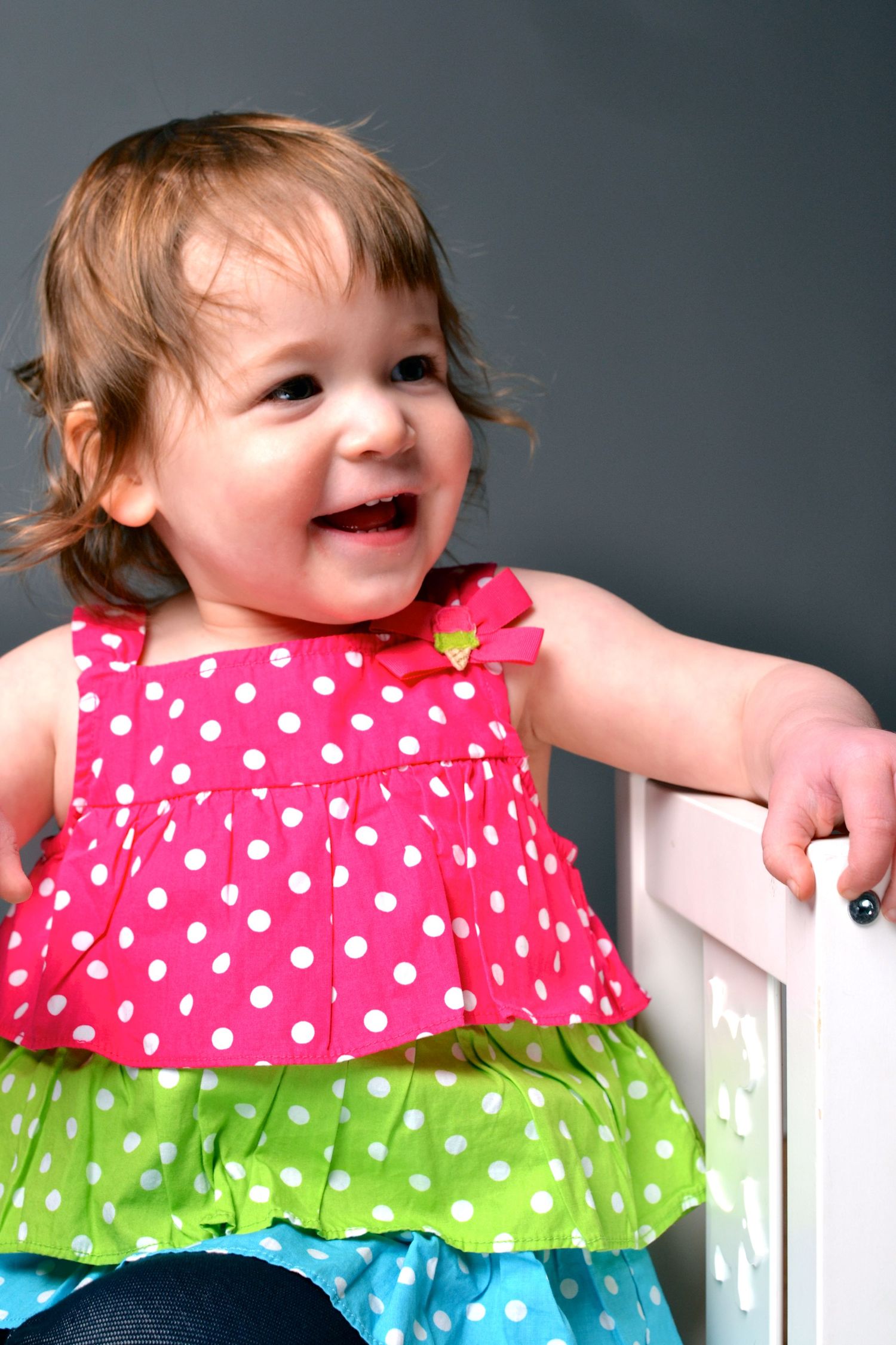 Smiling baby girl in colorful pink polka dot dress laughing joyfully while holding onto a white chair.