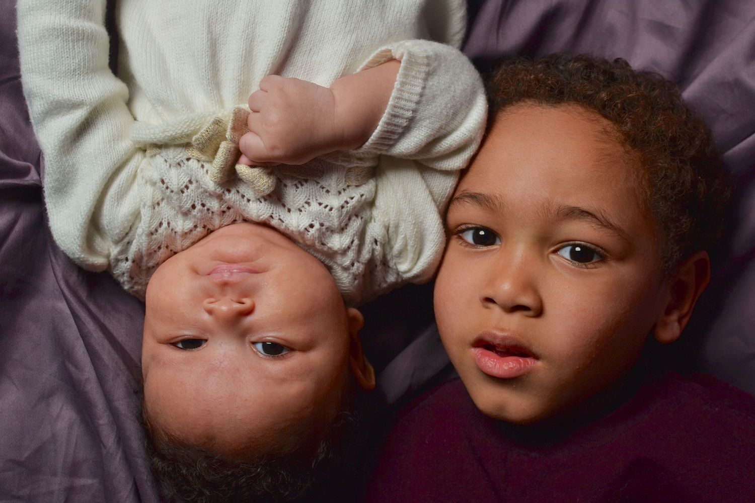 Two young children lying together, an infant upside down and a toddler boy looking at the camera on a purple blanket.