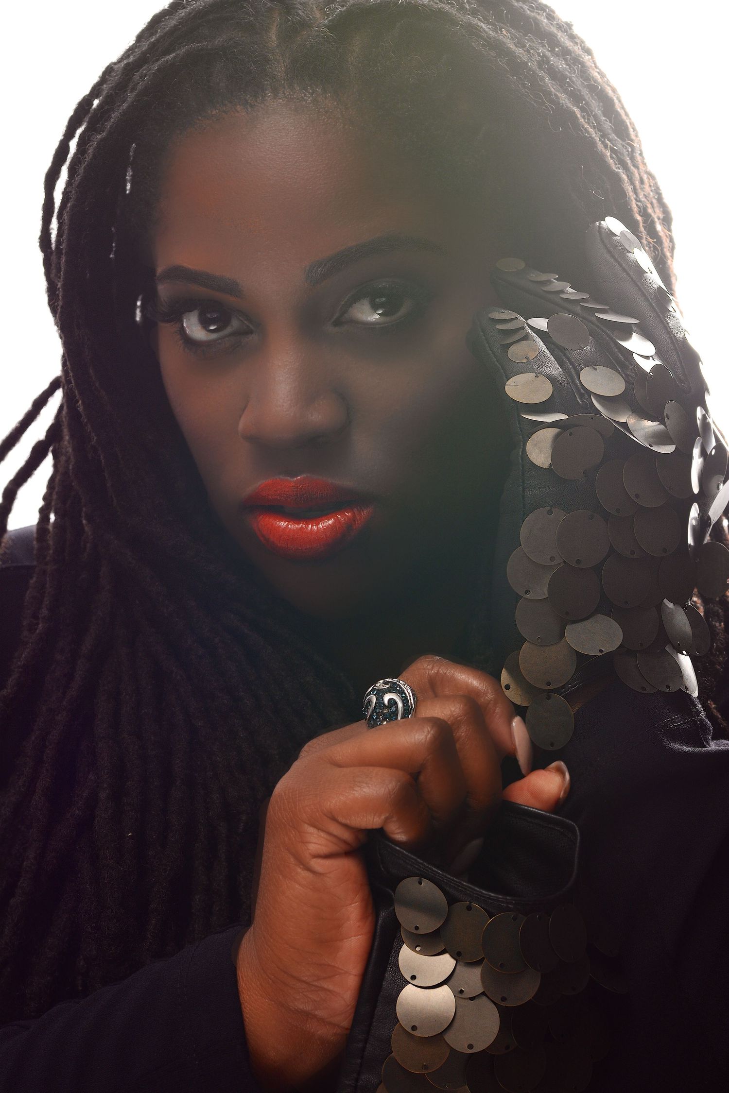 Dark-skinned Black woman with long braids and red lips holding sequined fabric in dramatic lighting.
