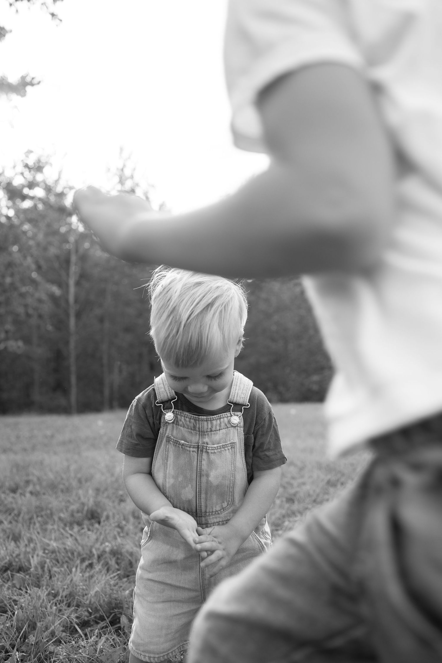 Black and white of boy looking at hands, candid.  Big brother blurred in front running.  Family photographer candid real moments