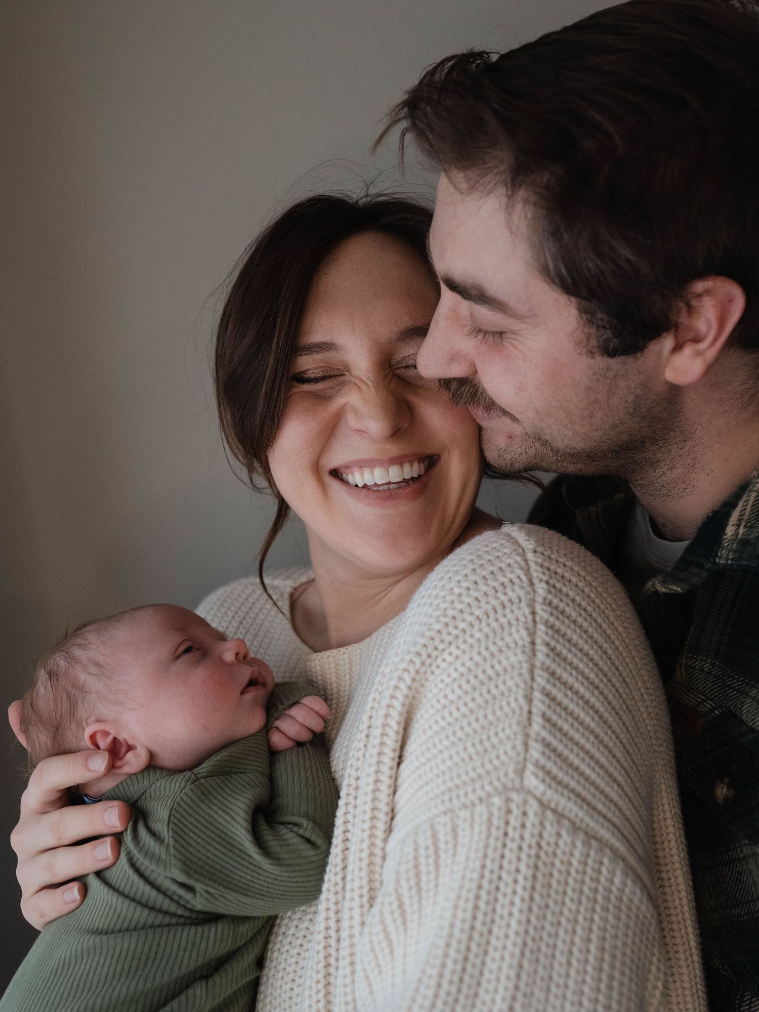 Couple in embrace holding newborn.  Father is standing behind mother, mother is smiling and facing camera.