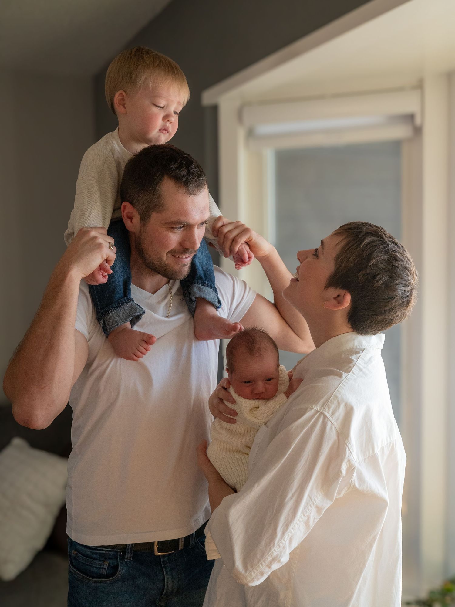 Colour image of family of 4, toddler is sitting on dads shoulders looking down at mom while she holds newborn.