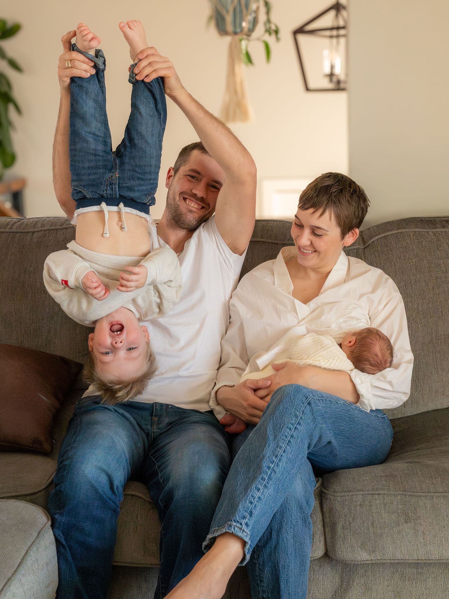 Colour image of family of 4 on sofa.  Father is holding toddler upside down while mother cradles newborn.