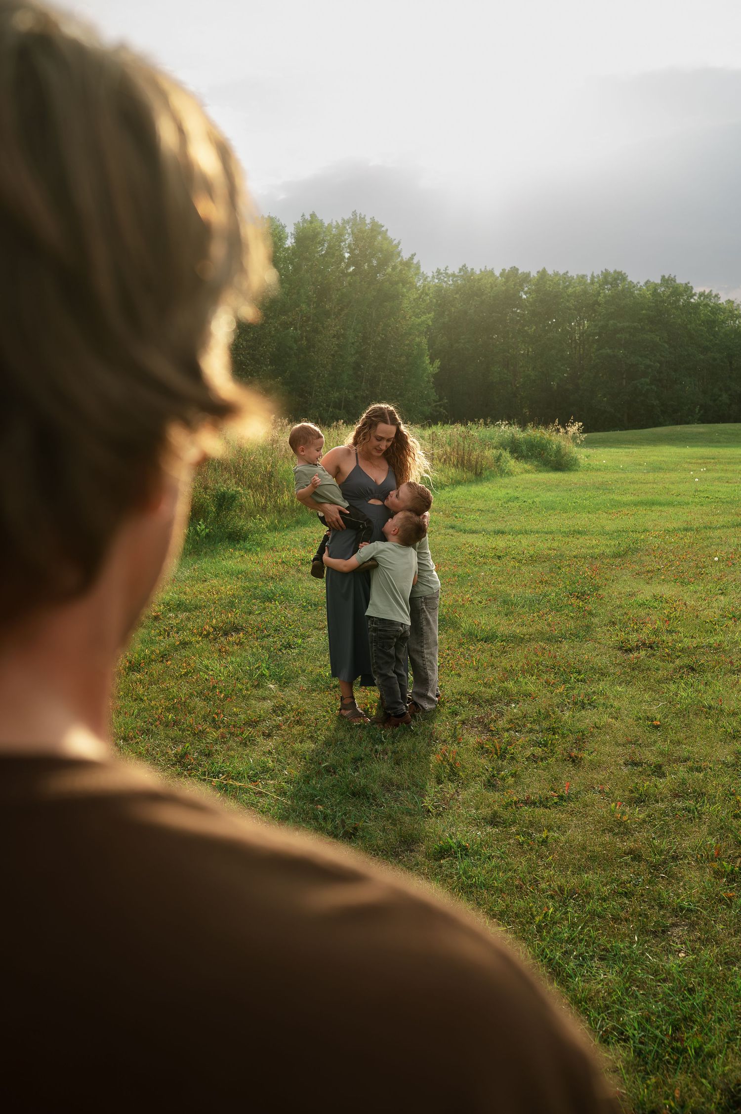 Family photograph looking over fathers shoulder at mom and children.  Golden hour, green field.  Lived in editing style