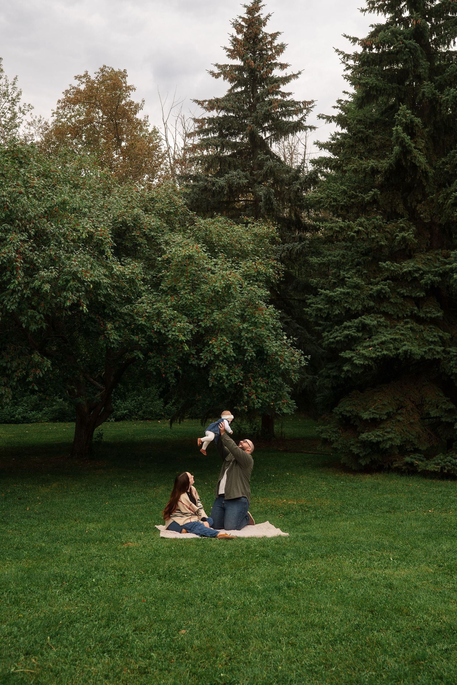 Documentary style photograph family of three, father holding daughter over head while mother looks up.  Taken in green park, trees, nature,