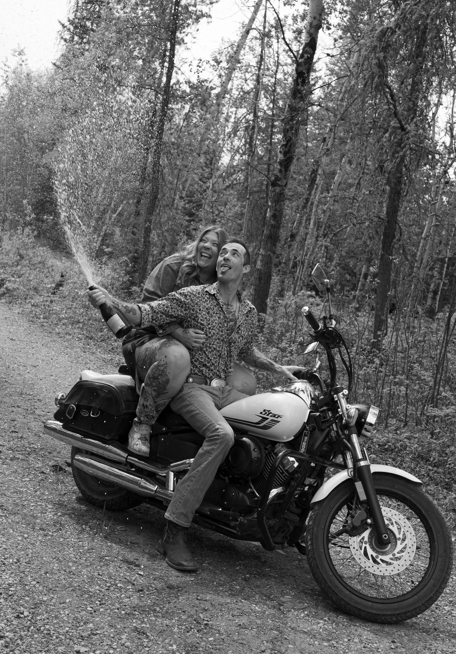 Black and white image of a couple goofily smiling on motorcycle during a Champagne pop and spray.