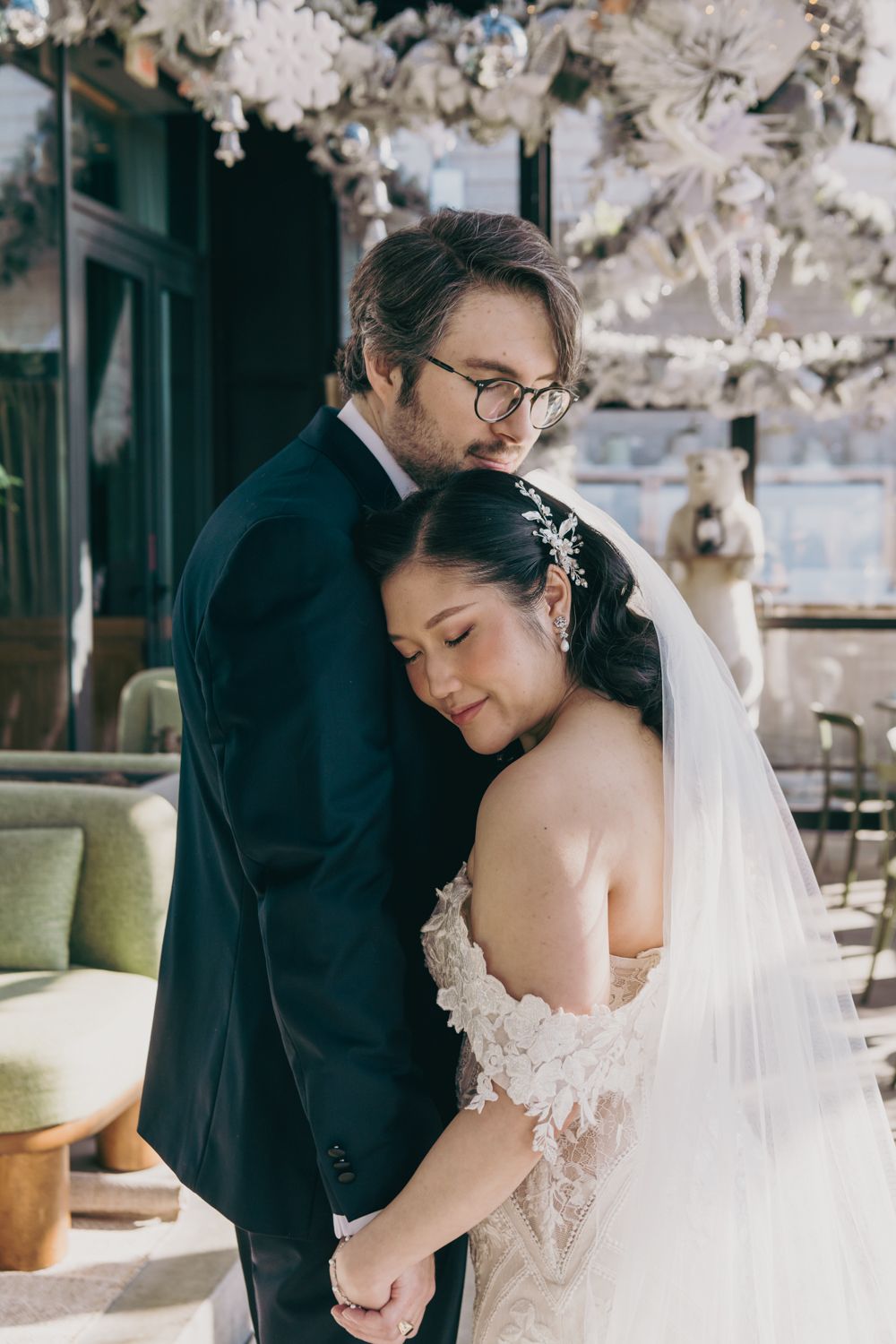 Bride in lace gown leans against groom in dark suit, sharing tender embrace under floral decorations.