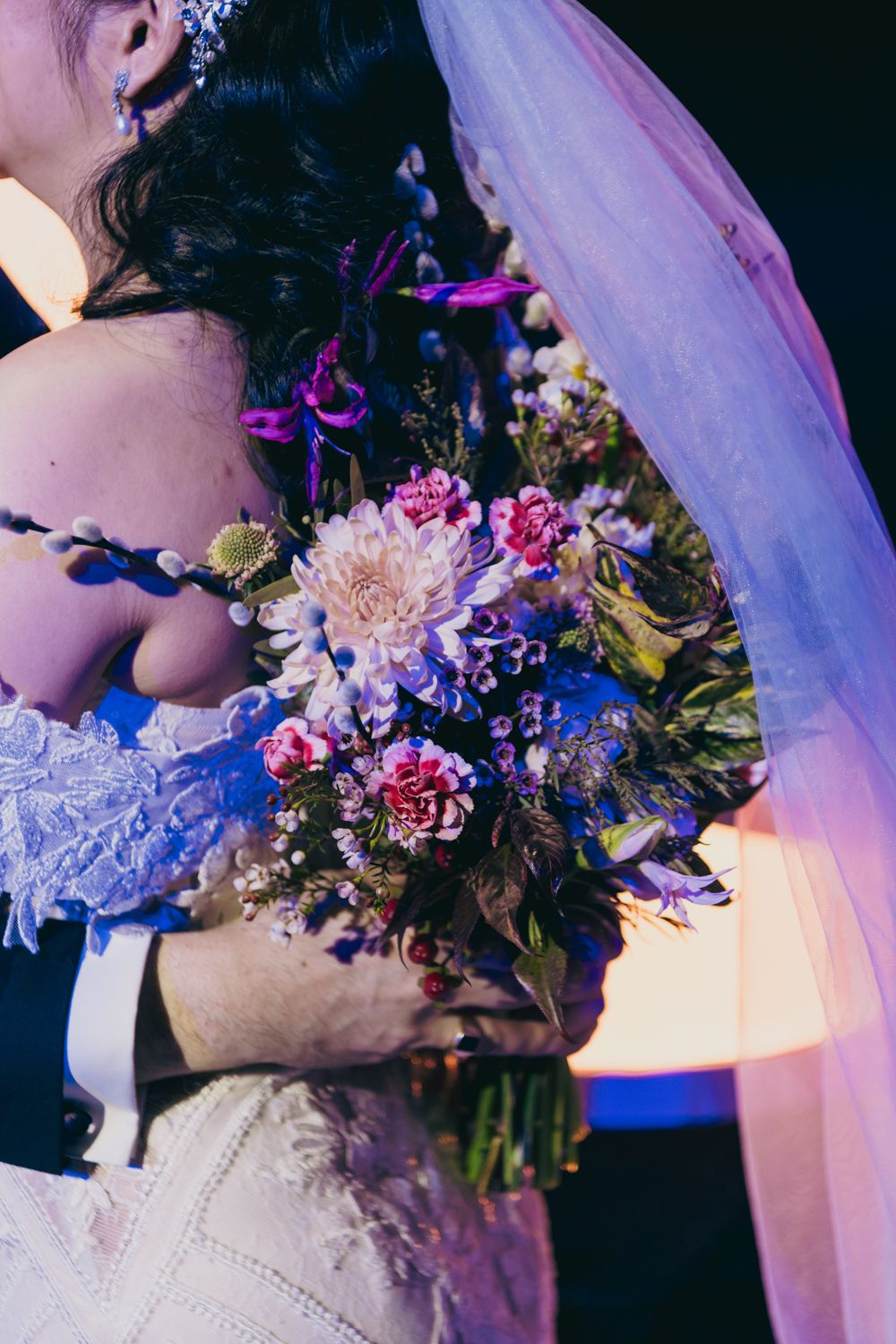 Bride holding a colorful wedding bouquet with pink dahlias and purple flowers while being embraced at her wedding reception.