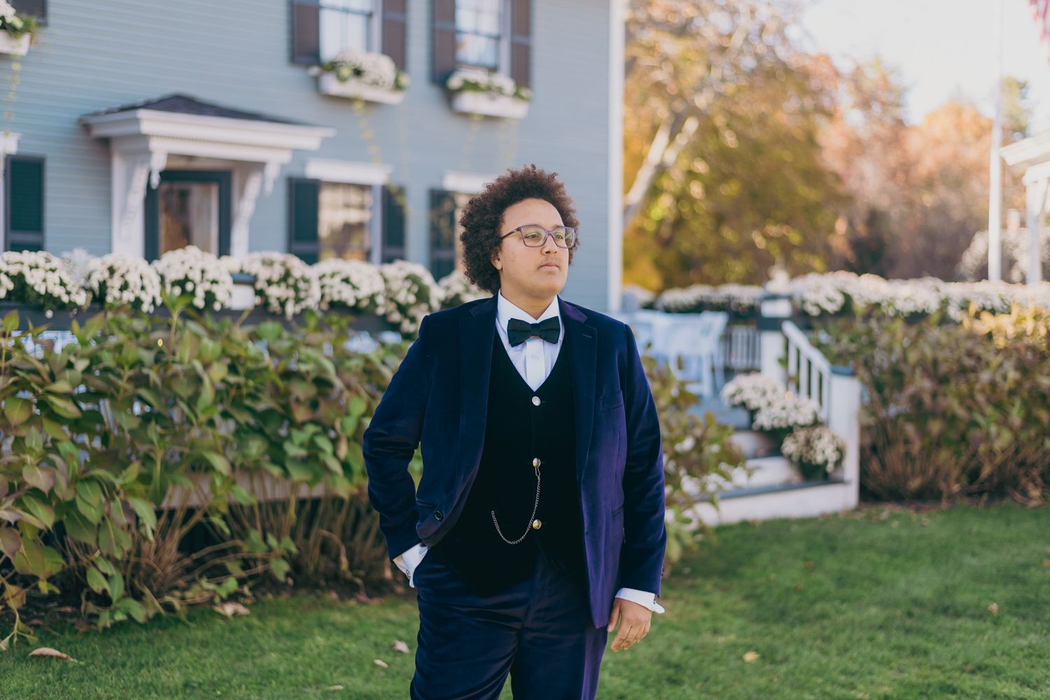 Man in navy blue velvet tuxedo with bow tie and pocket watch poses outdoors near white house with flowers.