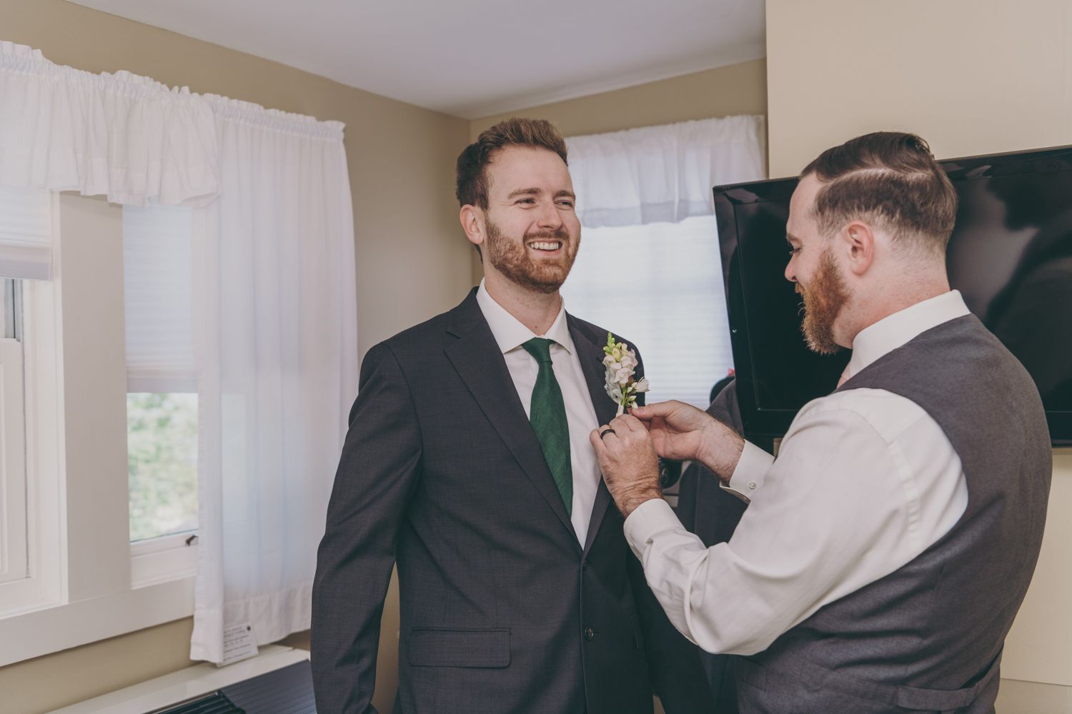 Smiling groom in grey suit receiving boutonniere from groomsman in vest before wedding.