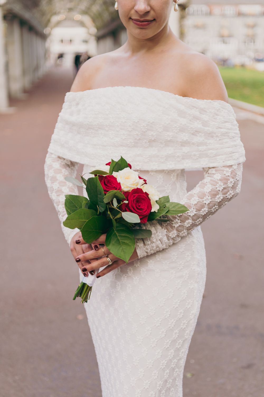 Bride in off-shoulder white lace gown holding red and white rose bouquet outdoors.