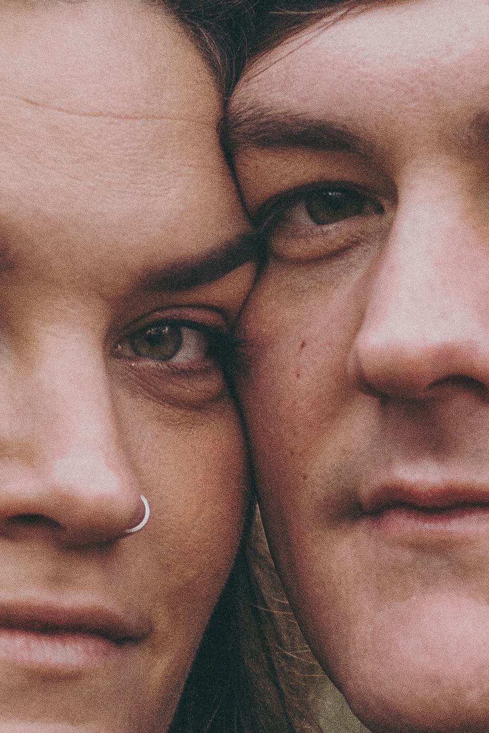 Close-up portrait of a couple with cheeks touching, woman with nose ring and man with brown hair gazing forward.
