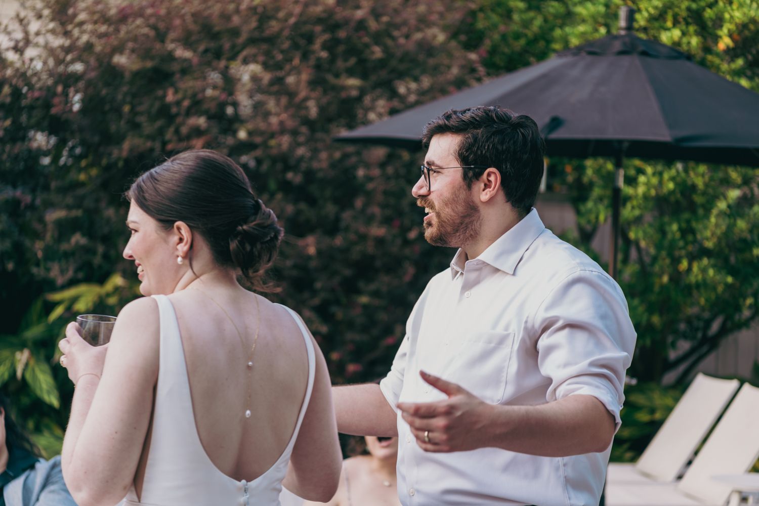 Bride and groom in casual attire enjoy conversation at an outdoor wedding reception surrounded by lush greenery.