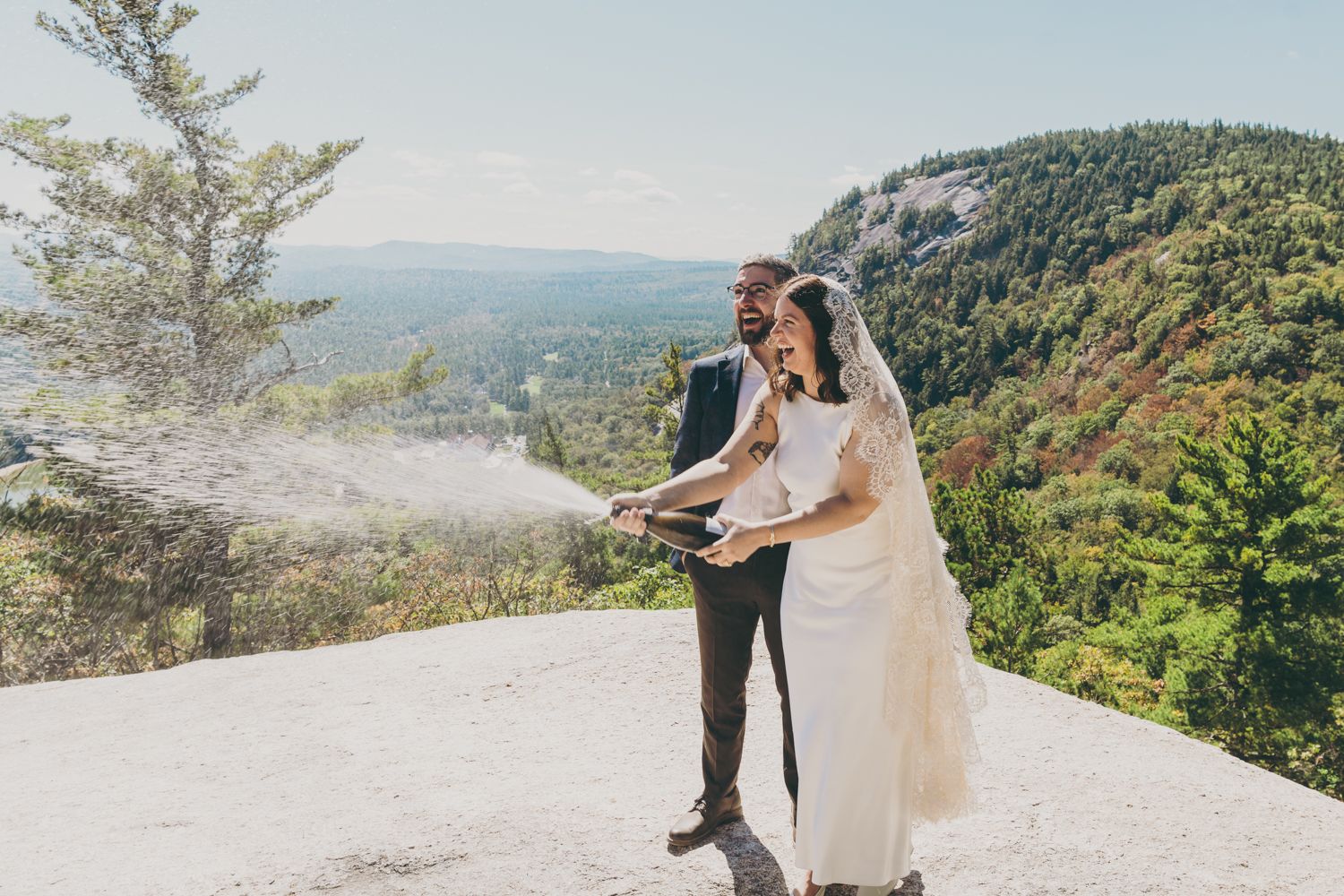 Joyful newlyweds pop champagne bottle on scenic rocky mountain overlook.