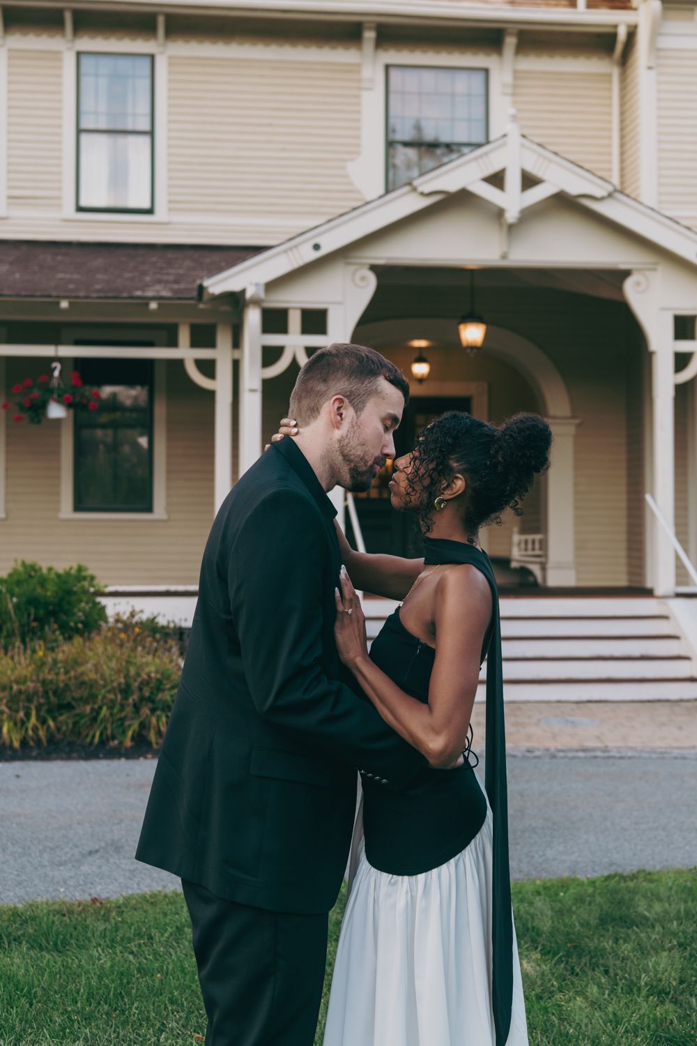 Interracial couple sharing a kiss in formal attire outside a charming yellow Victorian house.