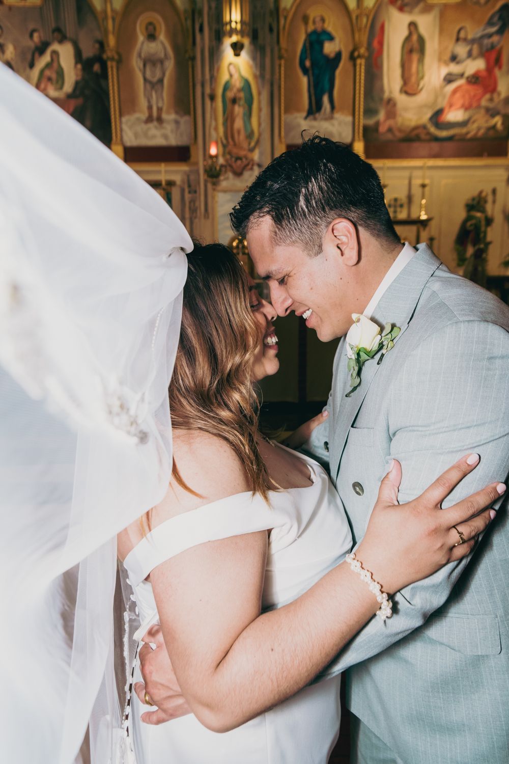 Bride and groom sharing an intimate moment under a veil inside an ornate church with religious iconography.