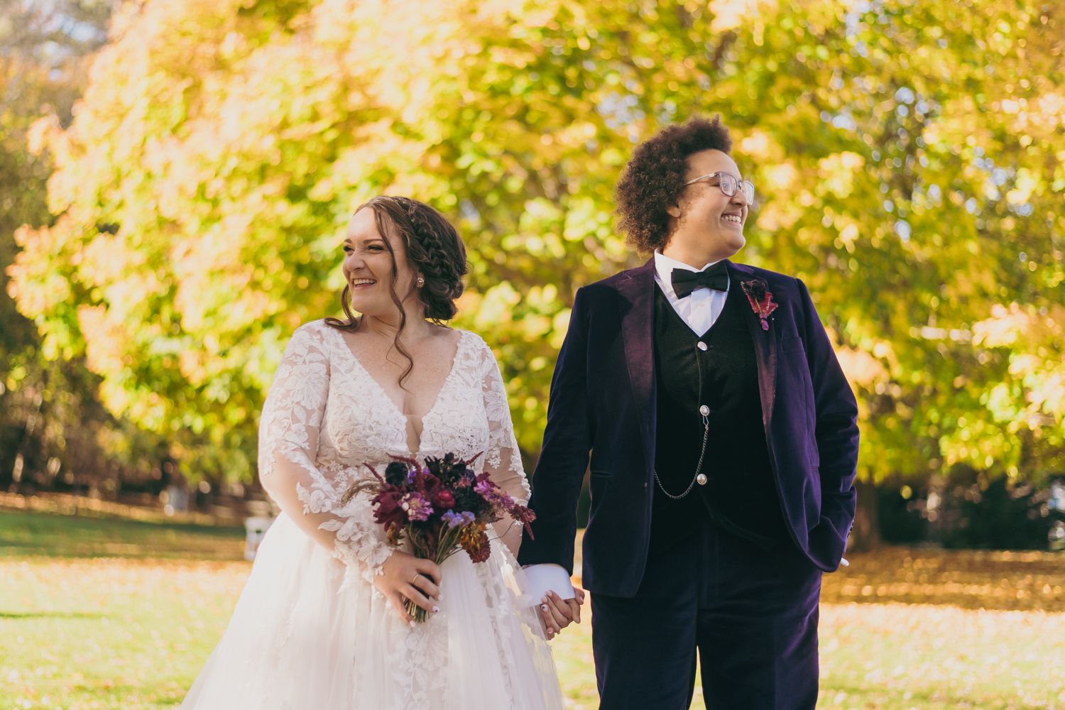 Bride and groom holding hands in front of vibrant yellow autumn trees outdoors.