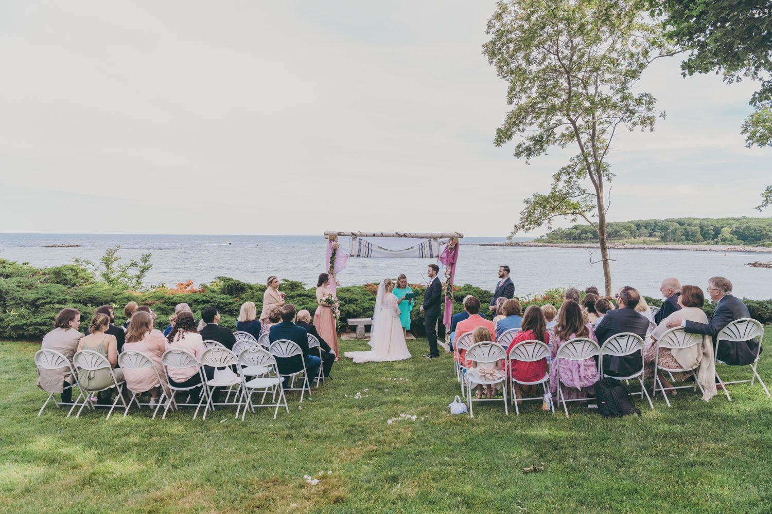 Intimate outdoor wedding ceremony with guests seated in white chairs facing floral arch by the water.