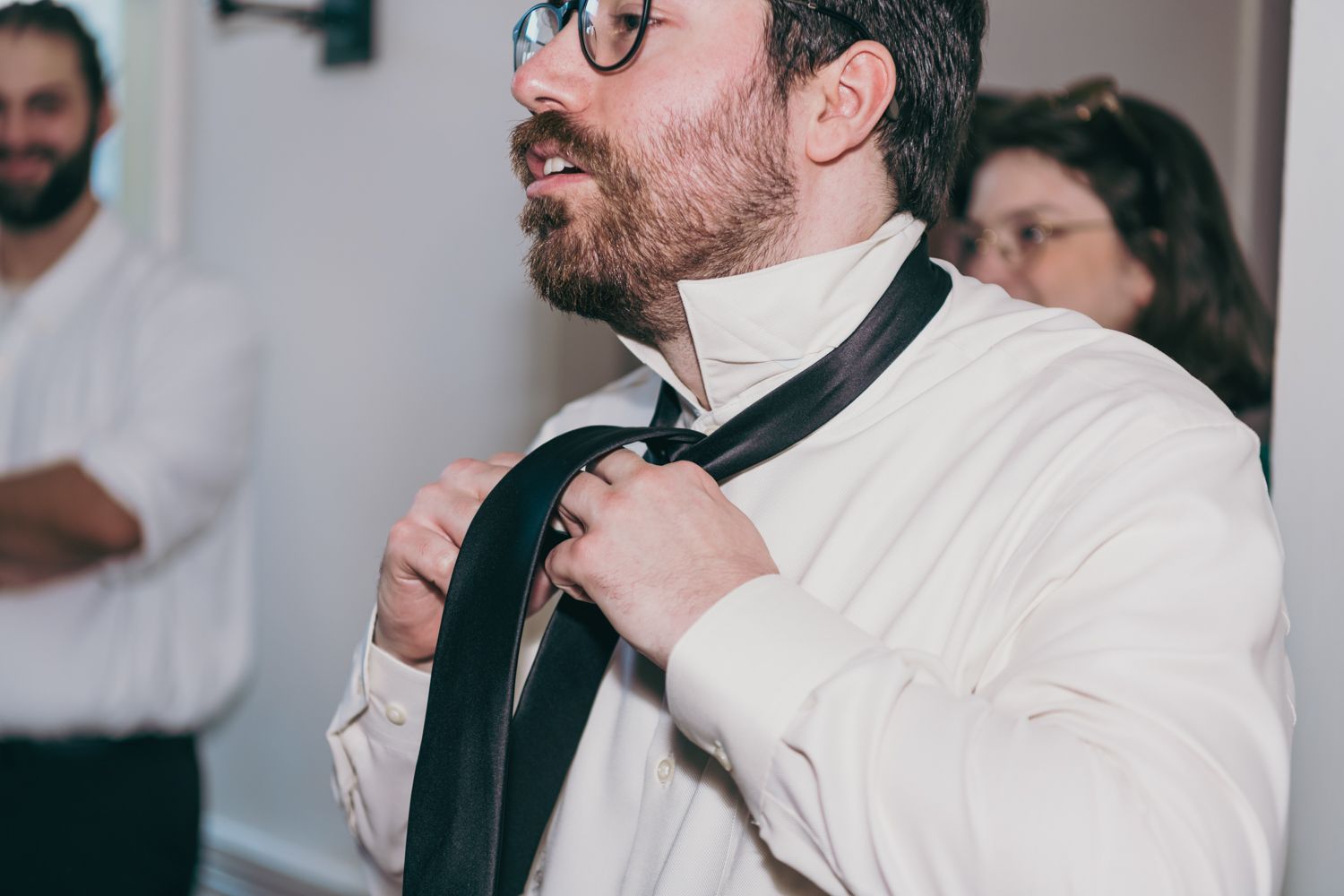 Groom with glasses and beard adjusting his black tie while getting ready for wedding.