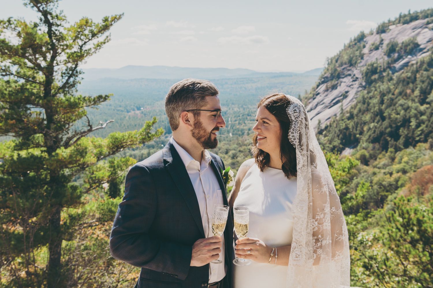 Smiling bride and groom toast with champagne against a mountain landscape.