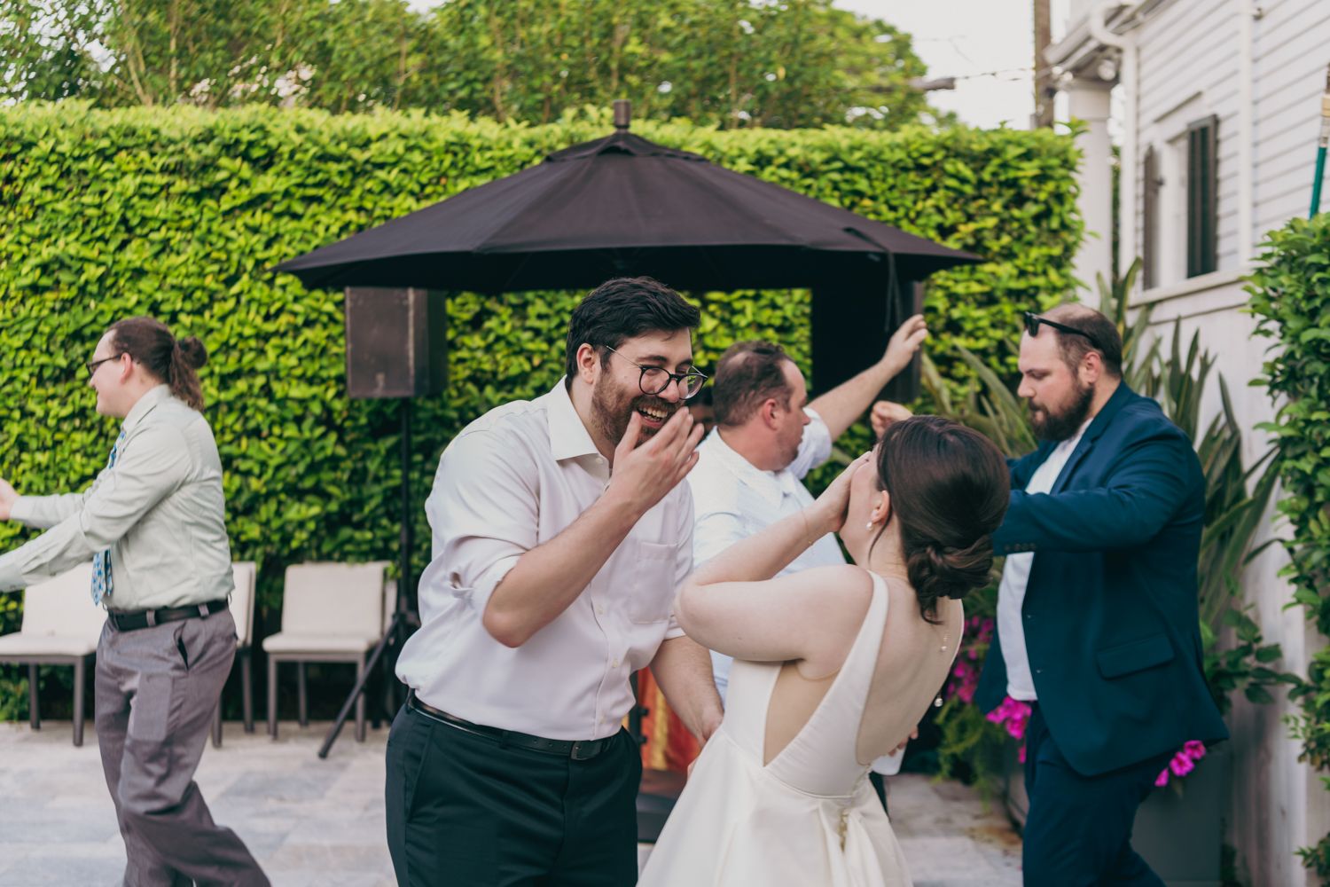 Wedding guests dance and celebrate together at an outdoor reception with green hedges and black umbrellas.