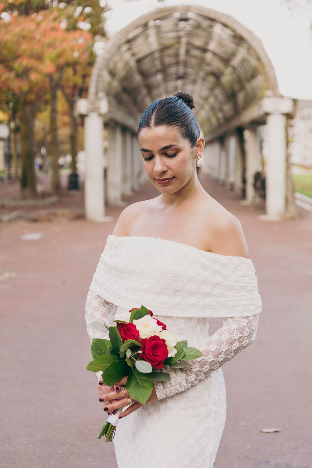 Bride in off-shoulder lace gown holding red and white rose bouquet under stone archway.