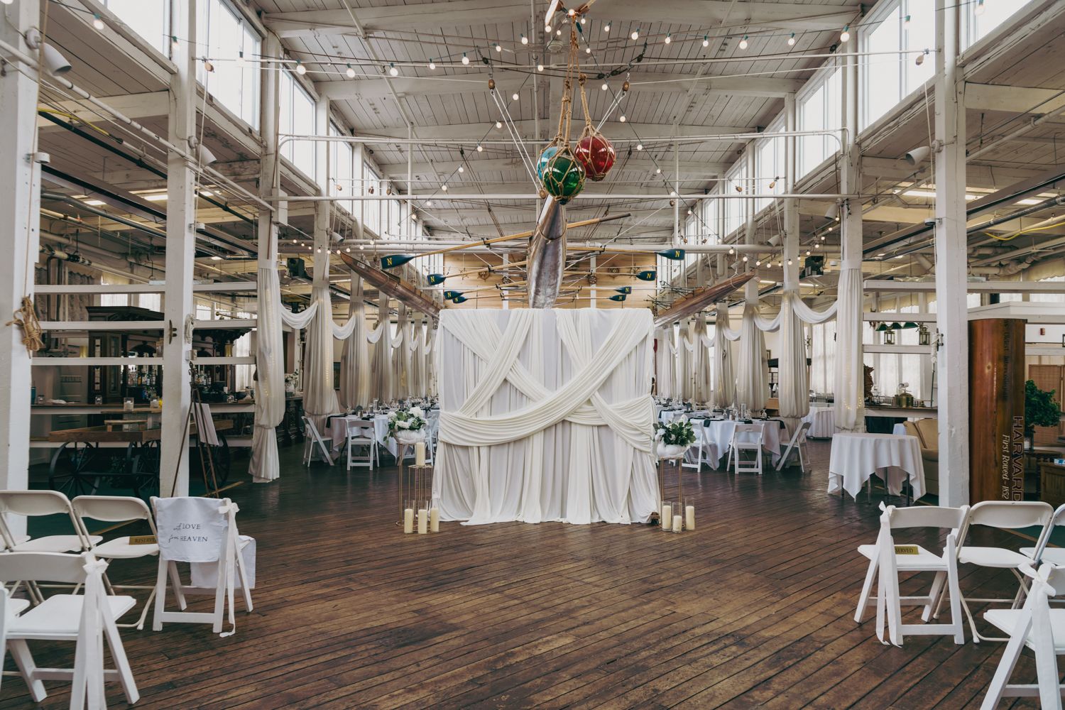 Elegant wedding ceremony setup in industrial venue with white draping, string lights, wooden floors, and white folding chairs.