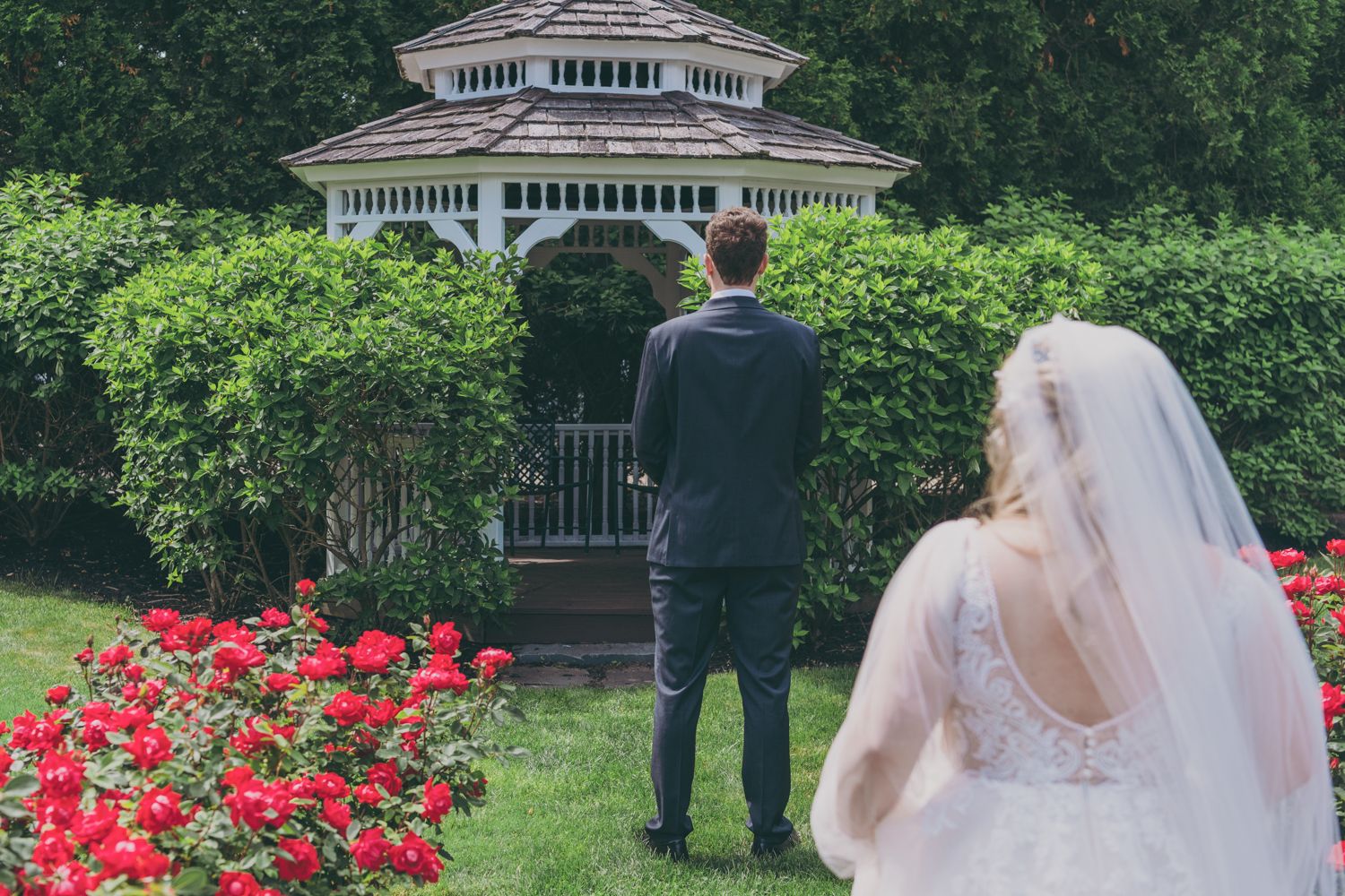 A bride in a white gown and veil approaches the groom near a gazebo surrounded by red roses.