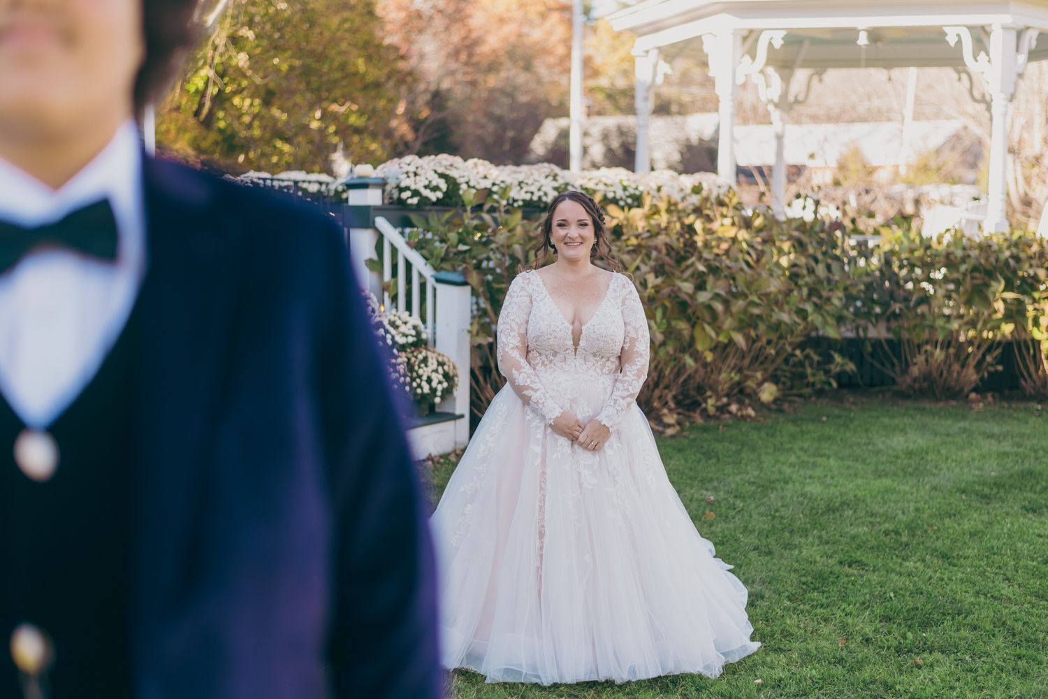 Bride in a long-sleeve lace ball gown smiling at groom near a floral gazebo outdoors.