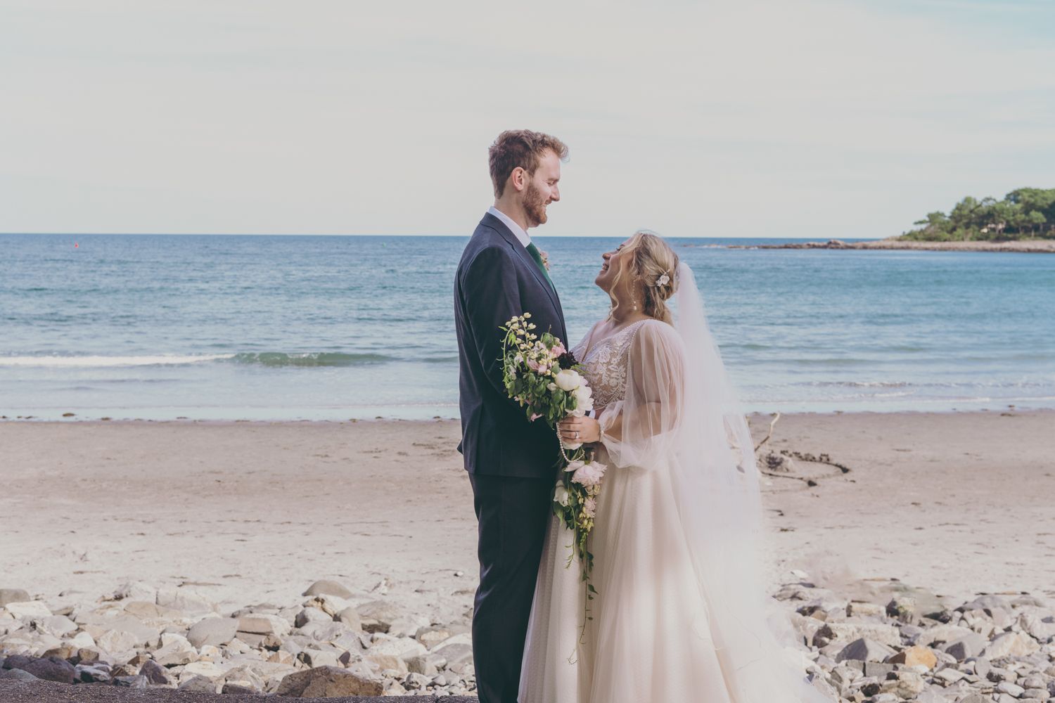 A bride and groom holding a bouquet stand facing each other on a sandy beach after their wedding.
