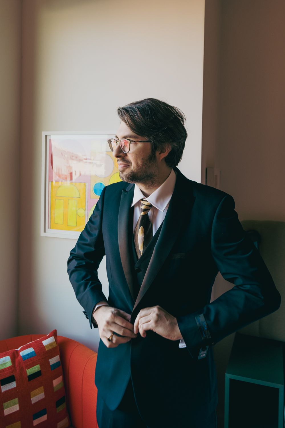 A groom in a dark suit and gold tie buttons his jacket while gazing to the side in a warmly decorated room.
